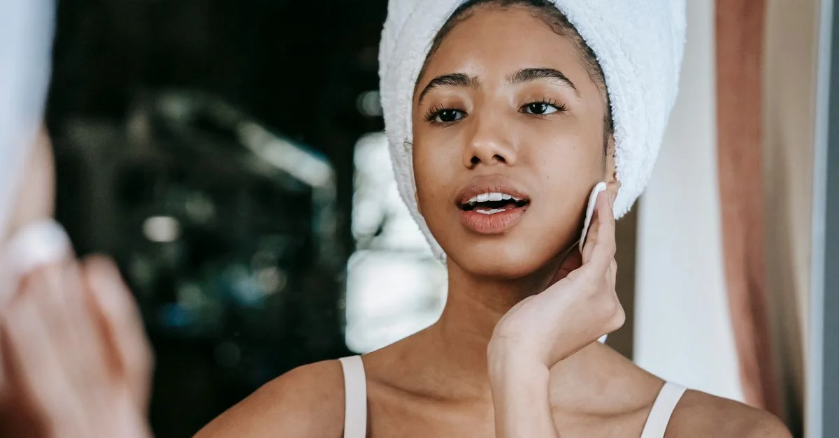 Une femme avec une serviette sur la tête utilise un coton pour nettoyer son visage, promouvant les soins de la peau et le bien-être.