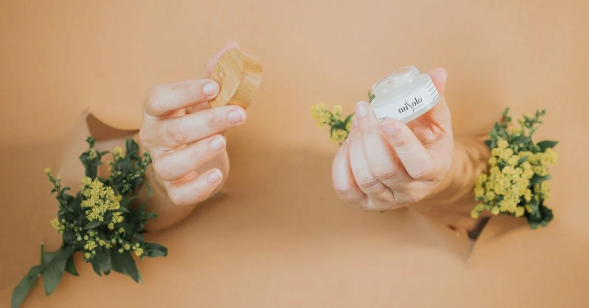 Une main tenant un pot de crème pour le visage parmi des fleurs jaunes sur un fond beige.