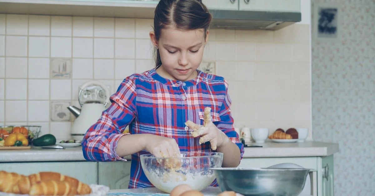 Enfant cuisinant dans une cuisine douillette, pétrissant de la pâte dans un bol, entourée d'ingrédients.